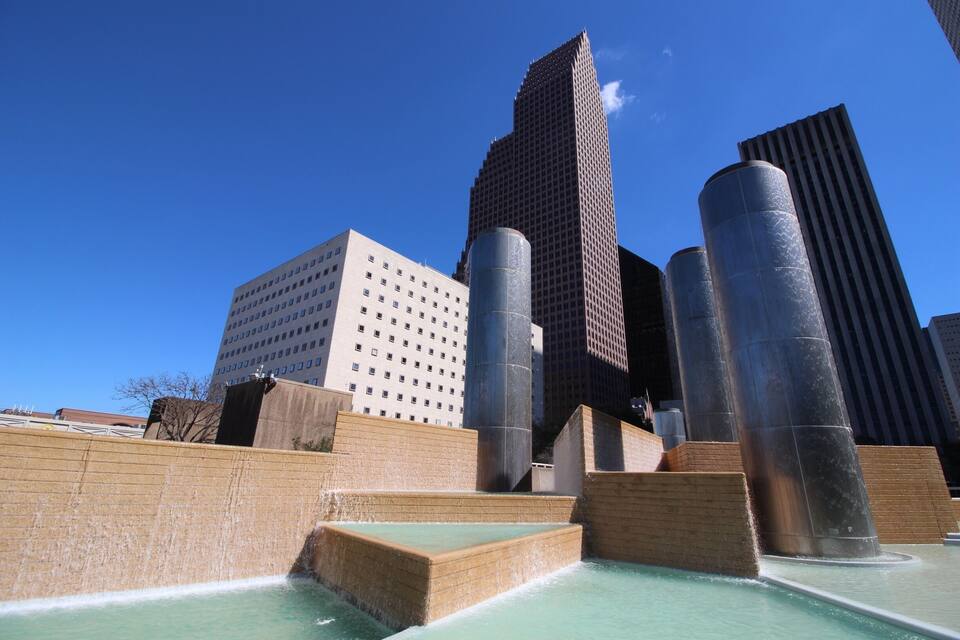 Un paisaje urbano moderno con edificios altos, una gran fuente de agua y un cielo azul despejado.
