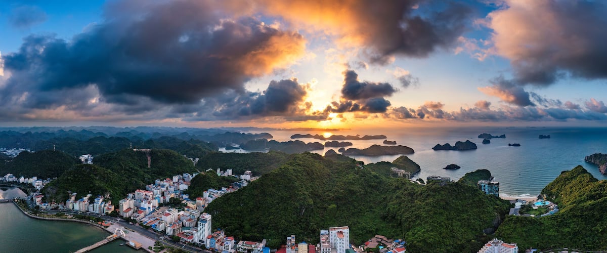 A coastal cityscape with dense greenery, a marina, and a sunset over the water.