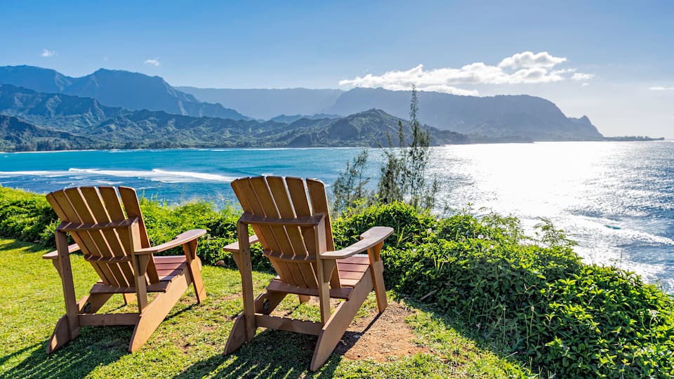 Duas cadeiras Adirondack de madeira com vista para a paisagem costeira e montanhas ao longe.