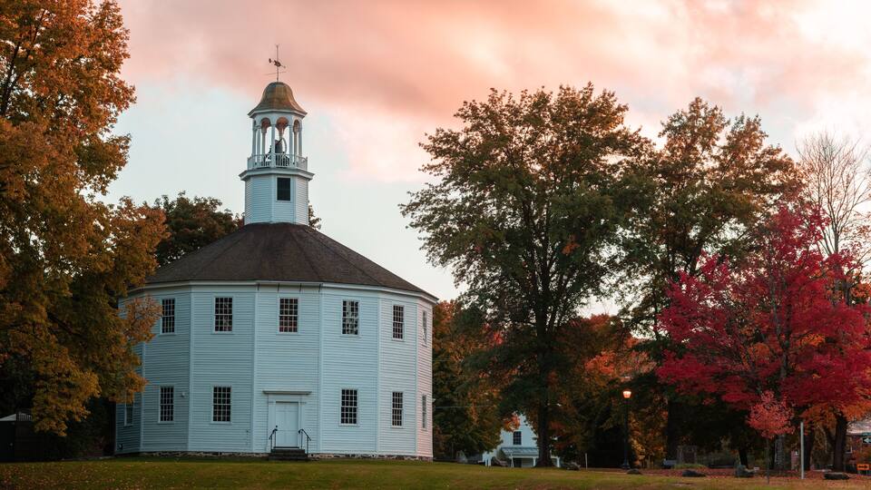 A white historic building with a bell tower, surrounded by autumn-colored trees.