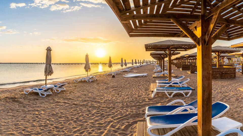A beach with sun loungers and umbrellas, a wooden pier extending into the sea, and a sunset in the background.