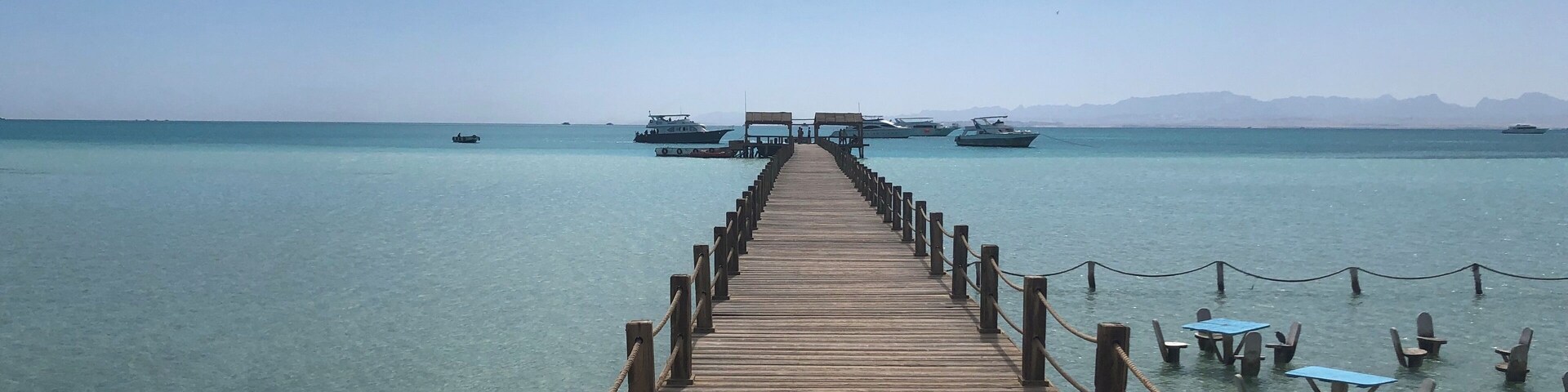 A wooden pier extending into clear turquoise water with boats anchored in the distance.
