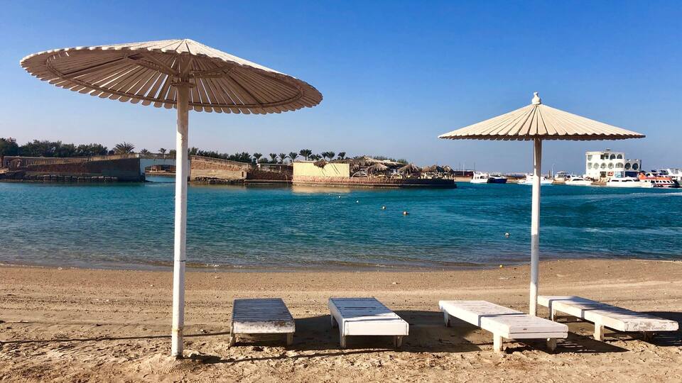 Una playa con sillas de descanso y sombrillas, un mar azul cristalino y una costa lejana con edificios y palmeras.