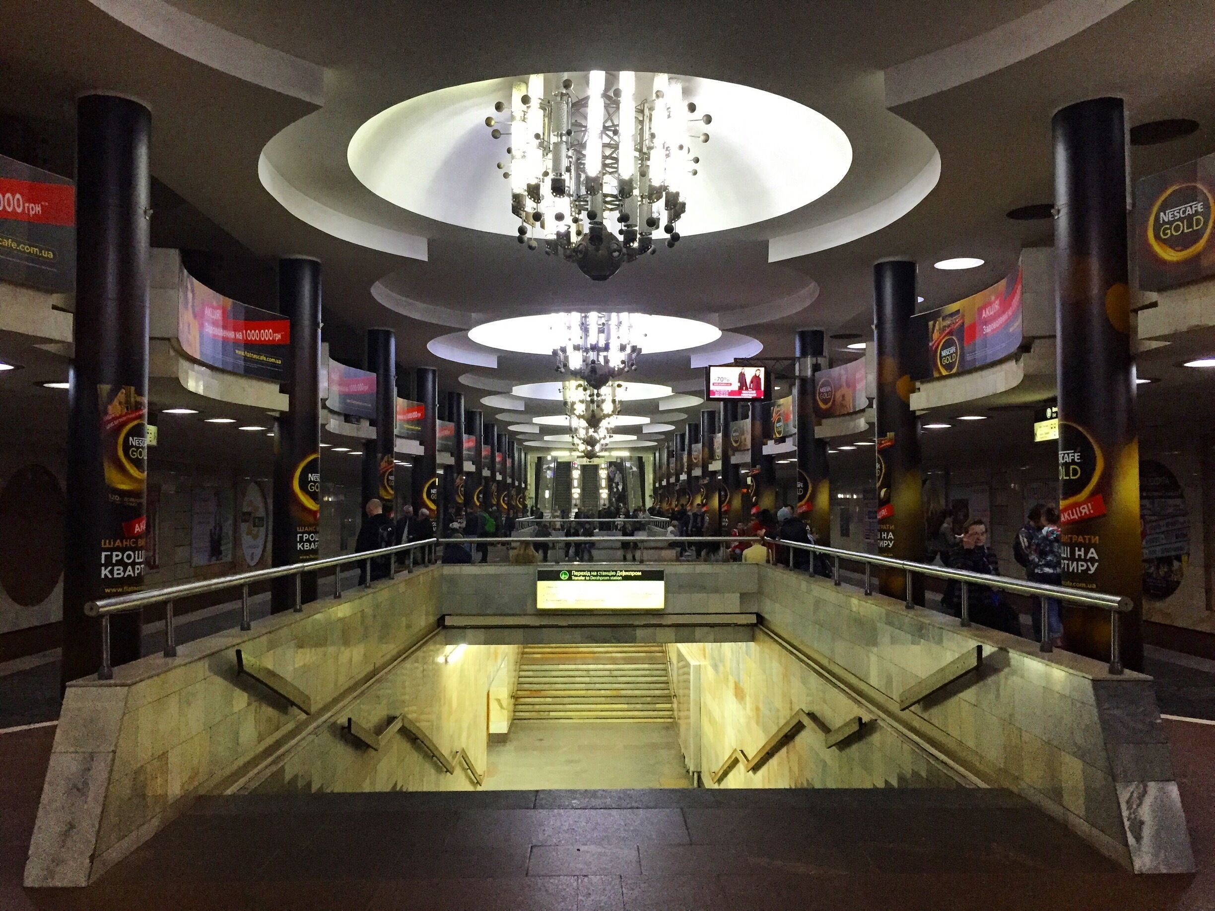 A modern indoor space with a central staircase, large chandeliers, and advertisements for Nescafe Gold.