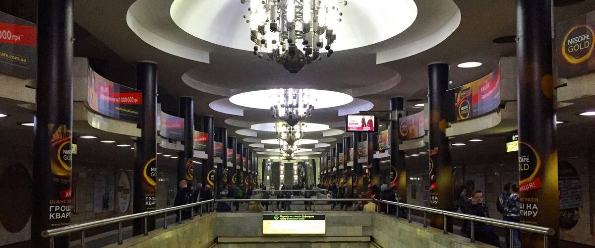 A modern indoor space with a central staircase, large chandeliers, and advertisements for Nescafe Gold.