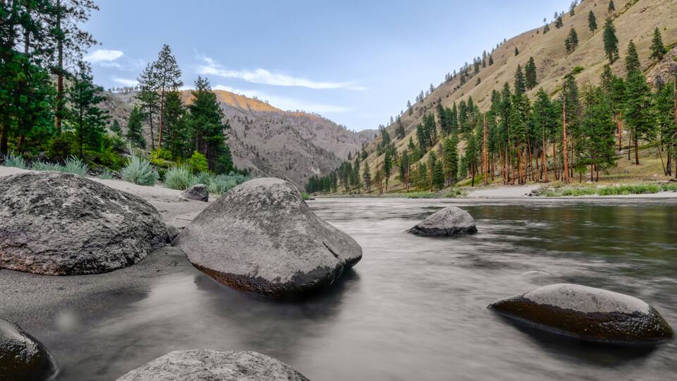 A river with large boulders, surrounded by a forest and hills.