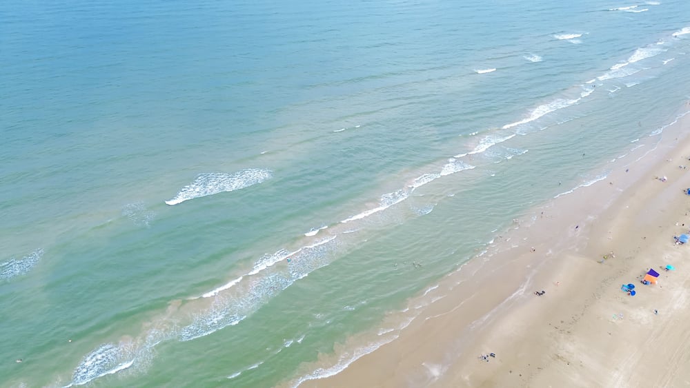 Luftaufnahme eines Strandes mit sanften Wellen, einem sandigen Ufer und vereinzelten Badegästen.