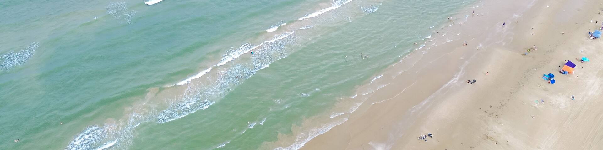 Diverse people enjoy beach activities swimming, surfing, sunbathing with umbrella, tent, camping and lounge chairs along sandy shoreline of South Beach in tropical Padre Island, Texas, aerial