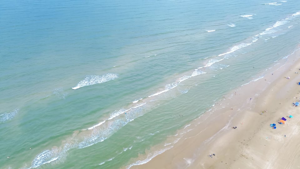 An aerial view of a beach with gentle waves, a sandy shore, and scattered beachgoers.