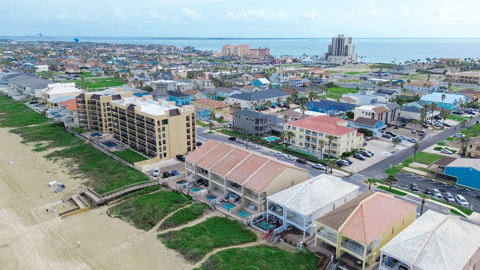 An aerial view of a coastal town with a beach, residential buildings, and a large hotel with a swimming pool.