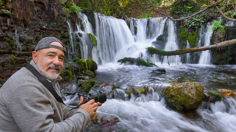 A waterfall with moss-covered rocks and a person taking a photo.