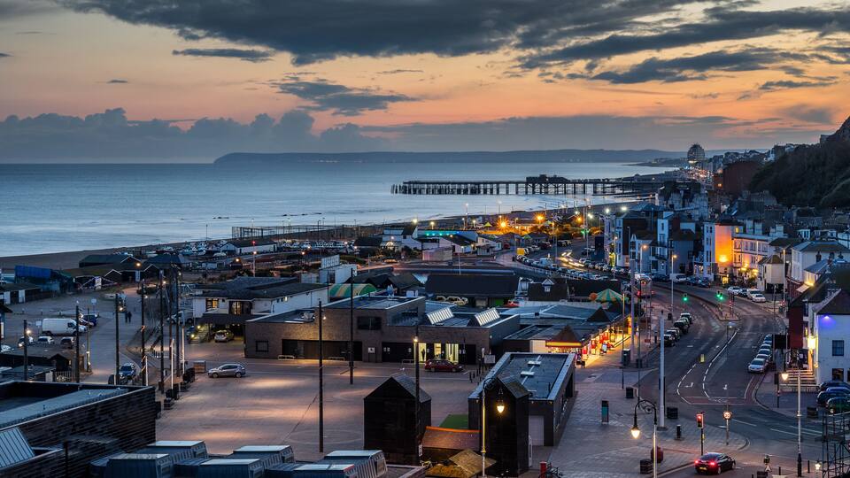 A coastal town at dusk with a pier extending into the sea, buildings lining the waterfront, and a road leading into the town.