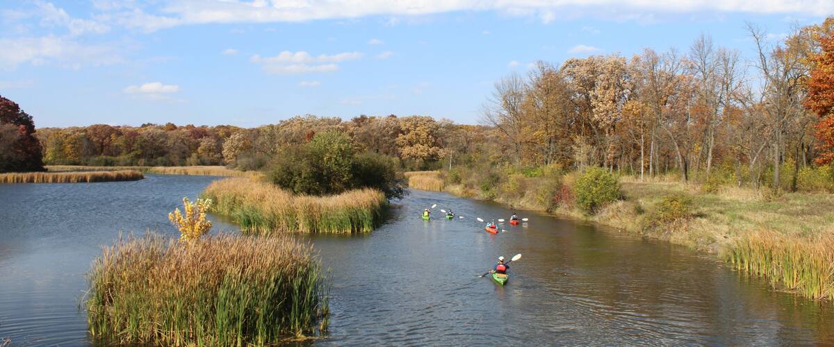A calm river with kayakers, surrounded by autumn foliage and a clear sky.