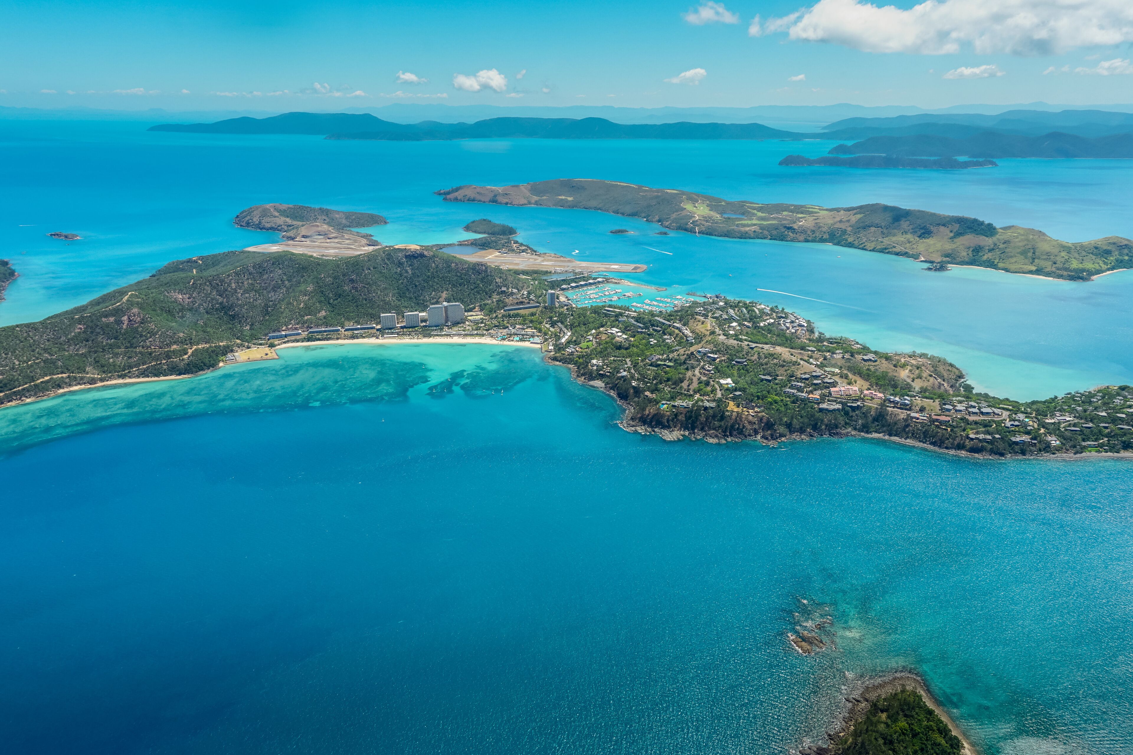 An aerial view of a coastal region with clear turquoise waters, a small island with dense vegetation, and a few buildings near the shore.