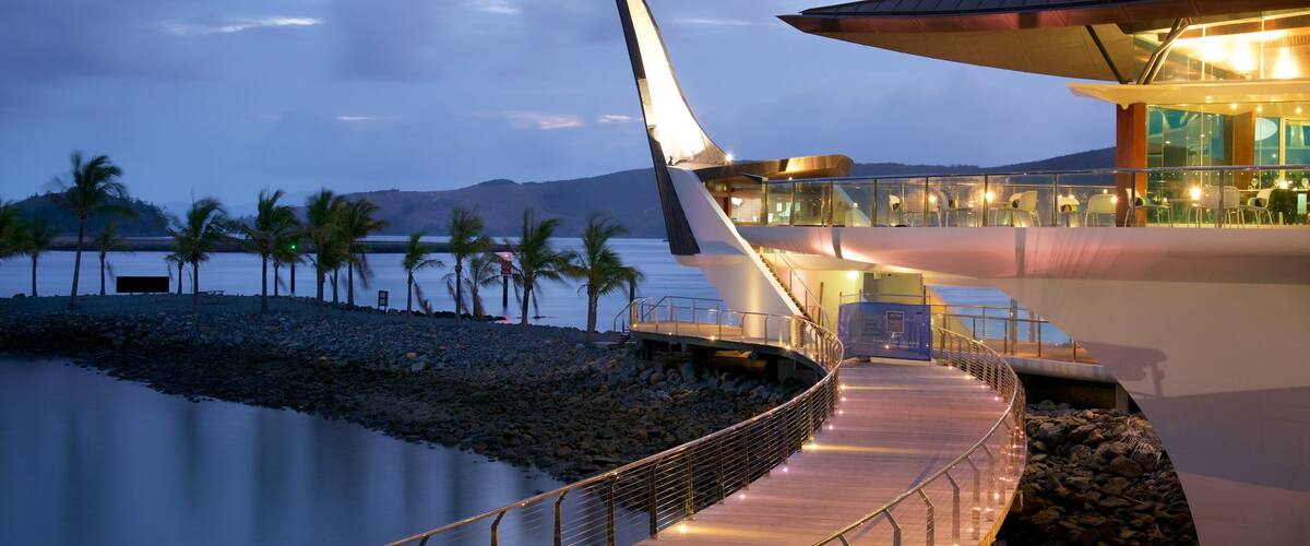 A waterfront resort with a modern building, palm trees, and a calm lake at dusk.