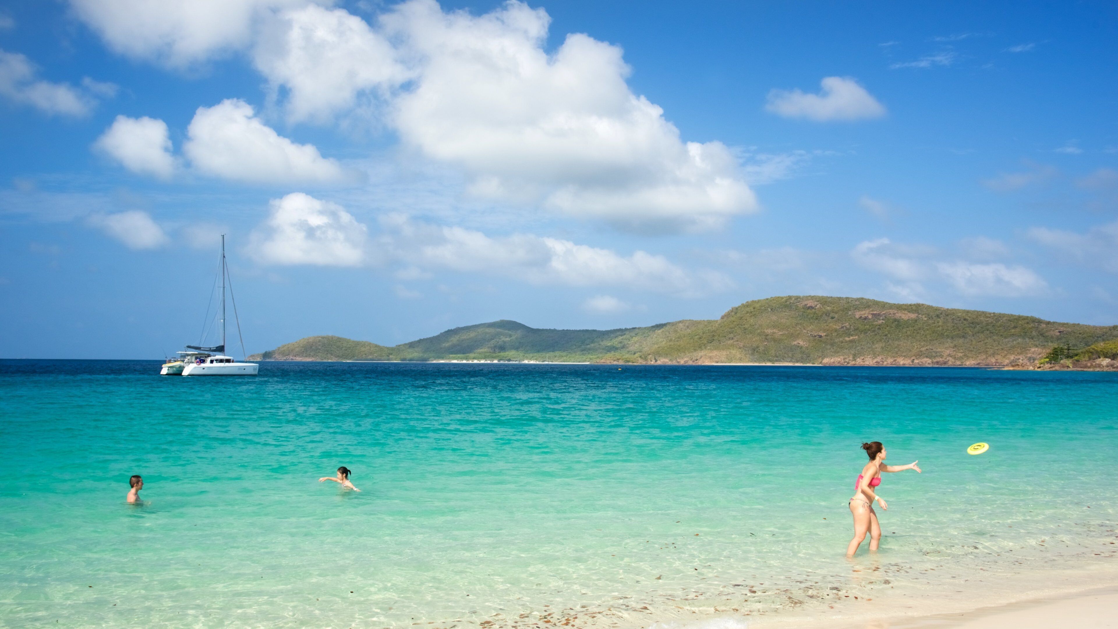 A beach with clear turquoise water, a sailboat, and a hilly landscape in the background.