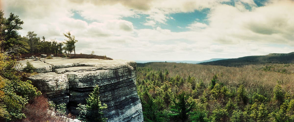Eine felsige Klippe mit vereinzelten Bäumen, ein bewaldetes Tal und in der Ferne Hügel unter einer teils bewölkten Himmel.