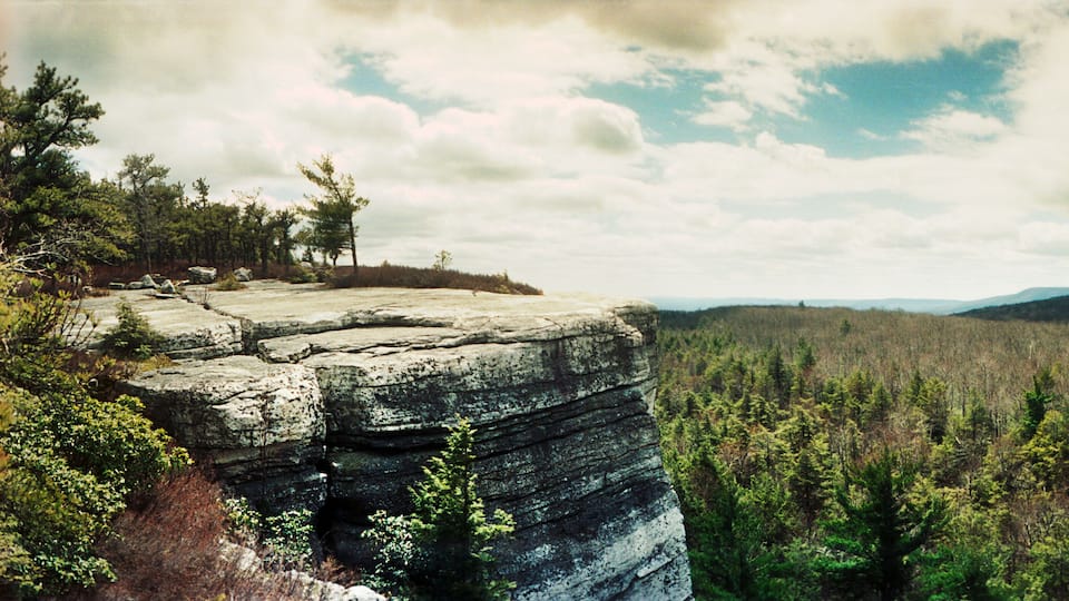 A rocky cliff with sparse trees, a forested valley, and distant hills under a partly cloudy sky.