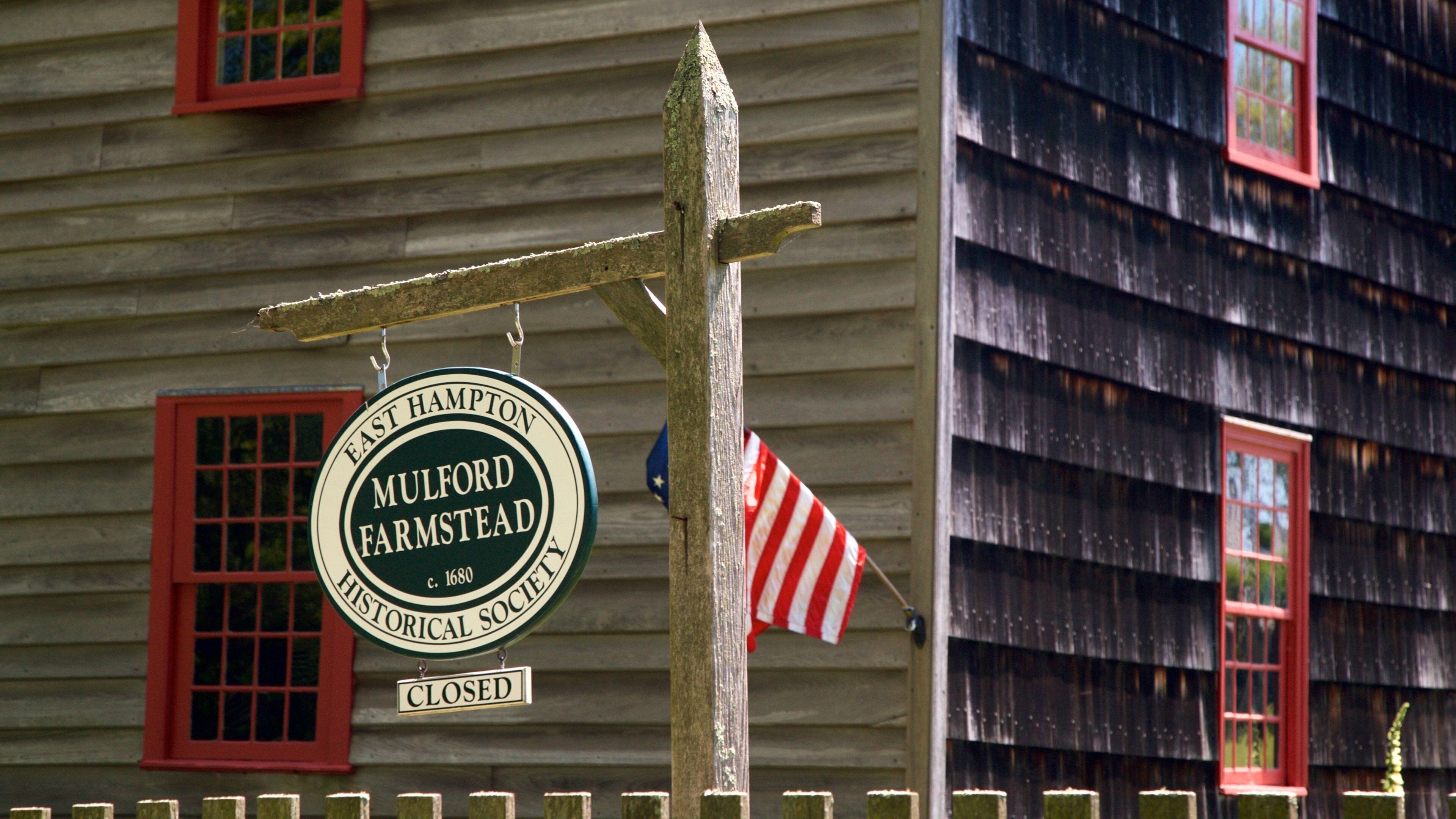 A closed sign for East Hampton Historical Society, Mulford Farmstead, with a red window and a wooden fence.