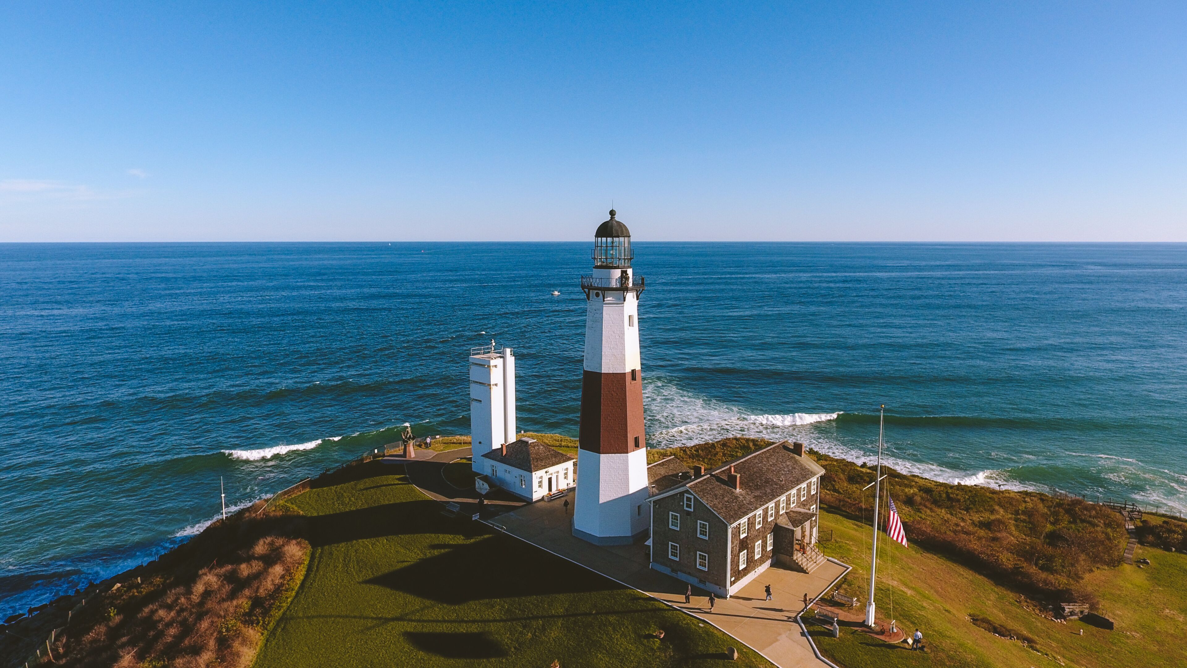 A lighthouse with a white and brown tower, a smaller white building, and a grassy area with a flagpole.