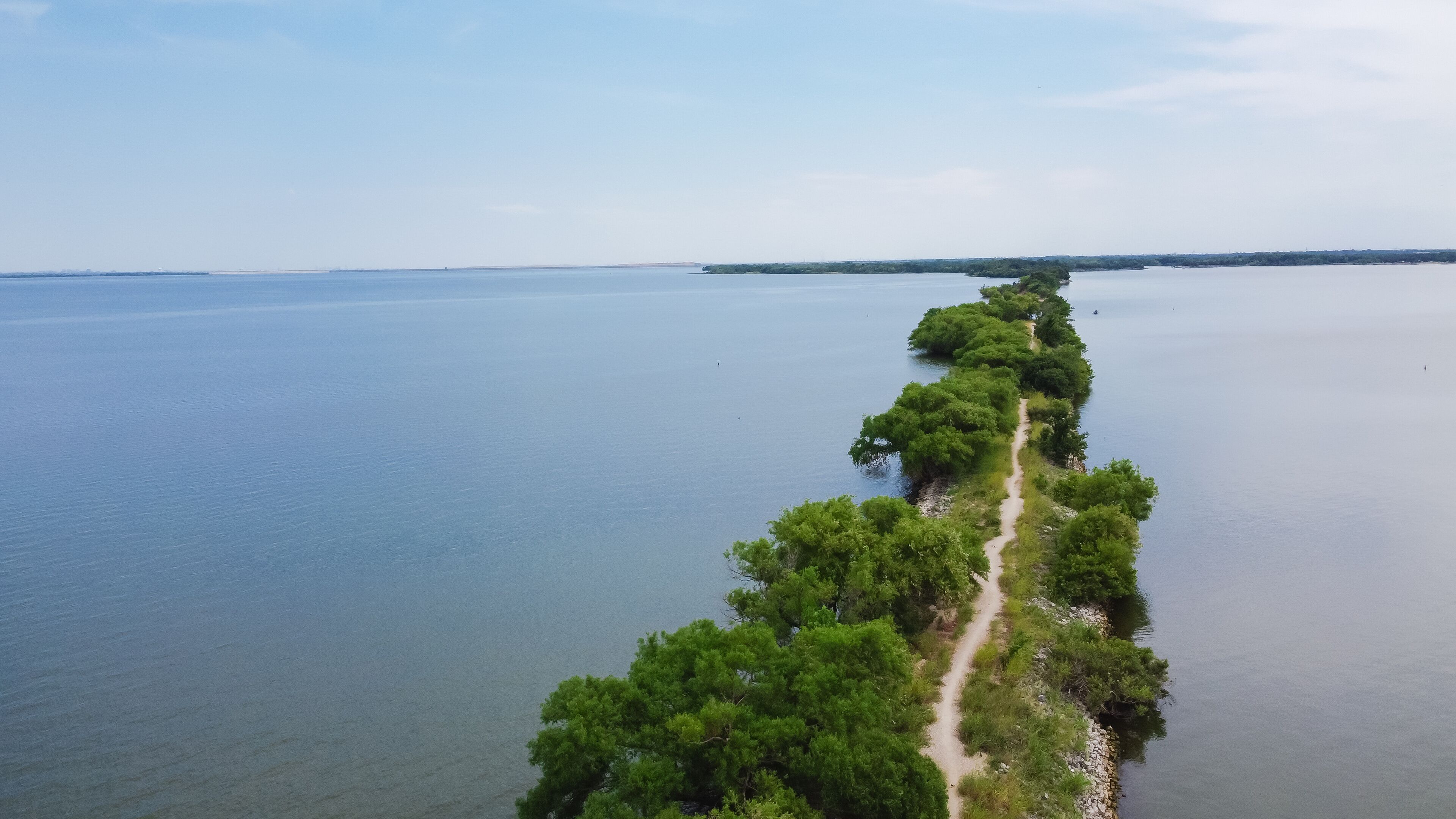 A long, narrow strip of land with dense green vegetation and a dirt path running through it, surrounded by calm water on both sides.