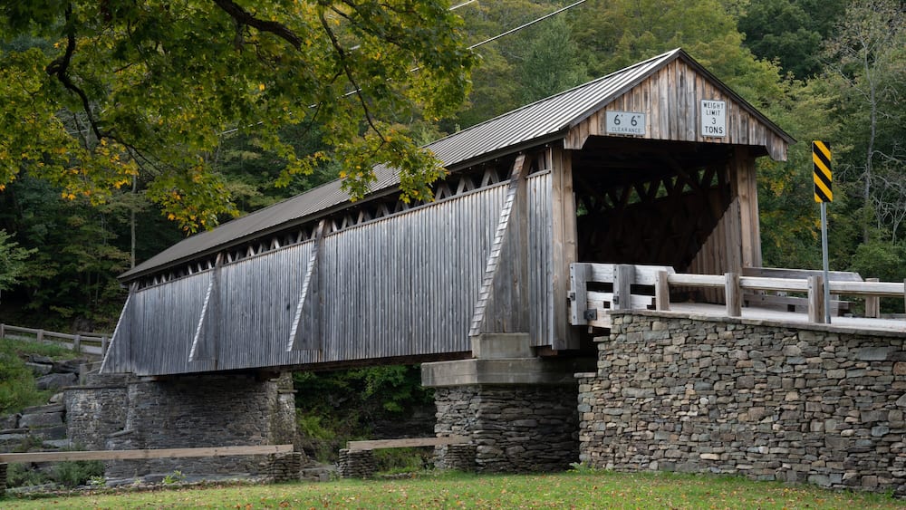 A wooden covered bridge with stone walls, surrounded by trees and a stone bridge in the background.
