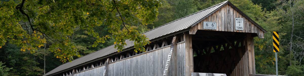 A wooden covered bridge with stone walls, surrounded by trees and a stone bridge in the background.