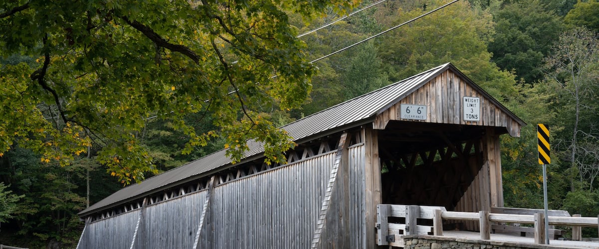 Un pont couvert en bois, avec des murs en pierre, entouré d’arbres et un pont en pierre en arrière-plan.