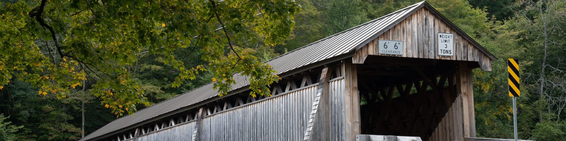 A wooden covered bridge with stone walls, surrounded by trees and a stone bridge in the background.