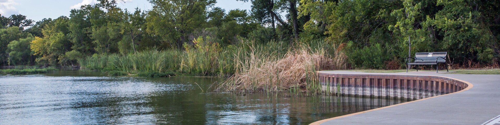 Uma passarela de concreto sinuosa à beira de um lago calmo, com árvores e um banco.