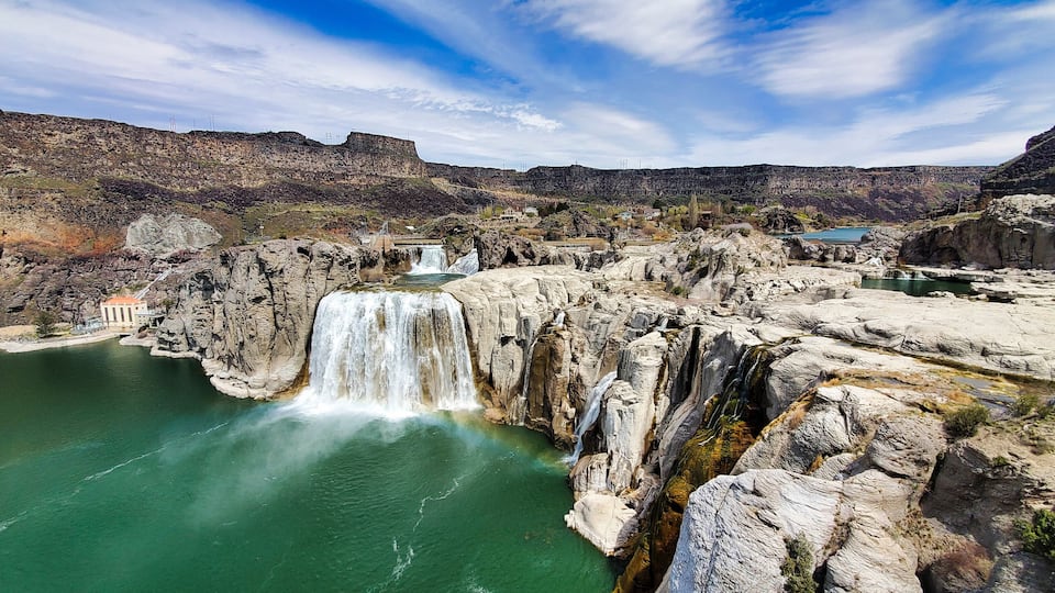 A large waterfall cascading into a turquoise lake surrounded by rocky cliffs and a small building.