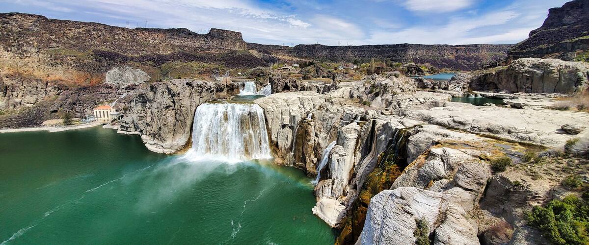 A large waterfall cascading into a turquoise lake surrounded by rocky cliffs and a small building.