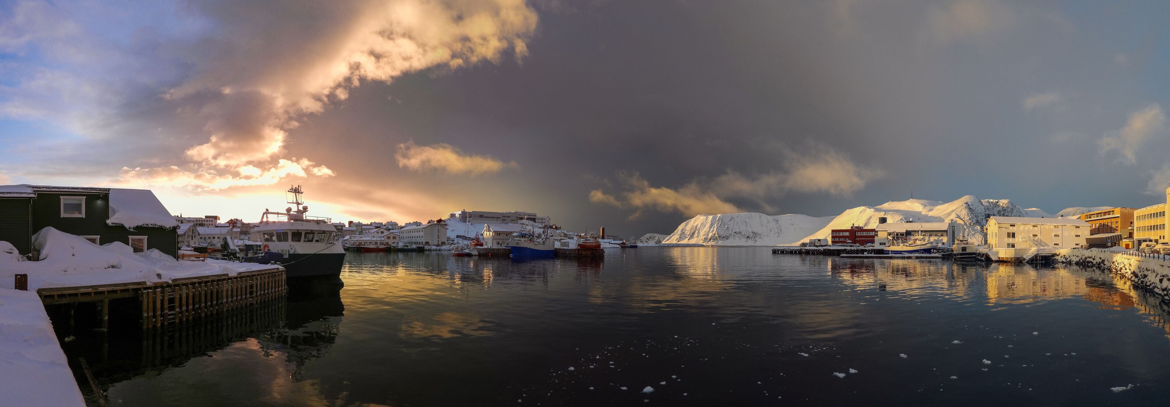 A snowy harbor with boats, buildings, and a mountain in the background.