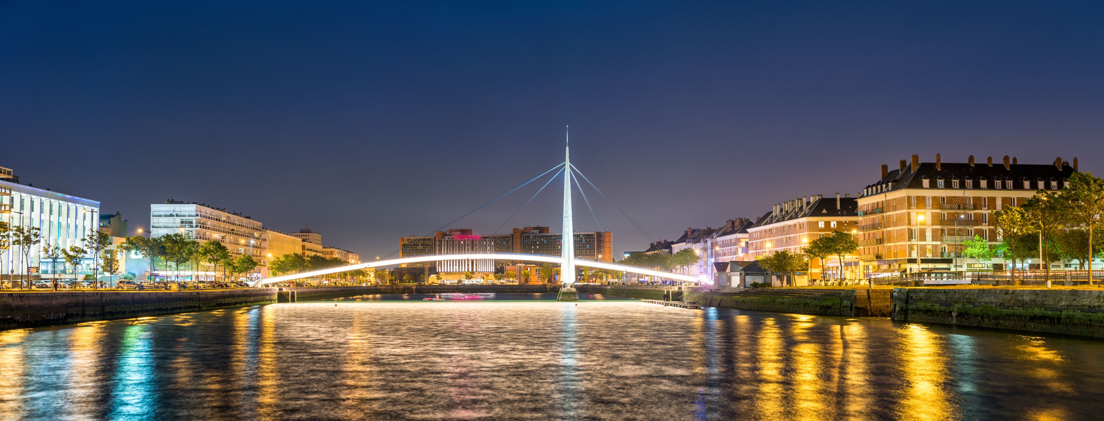 A nighttime cityscape with a prominent illuminated bridge over a river, reflecting city lights.