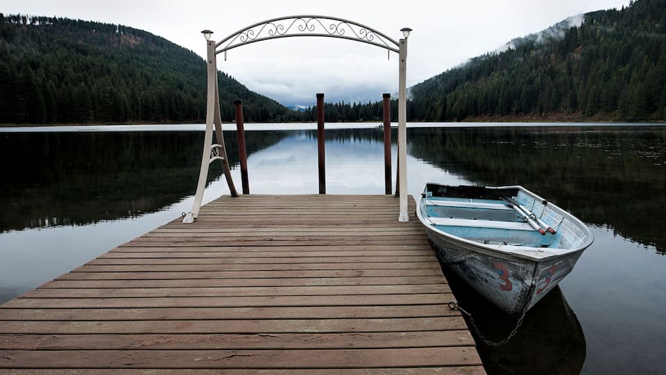 A wooden dock with a boat tied to it, surrounded by calm water and forested hills.
