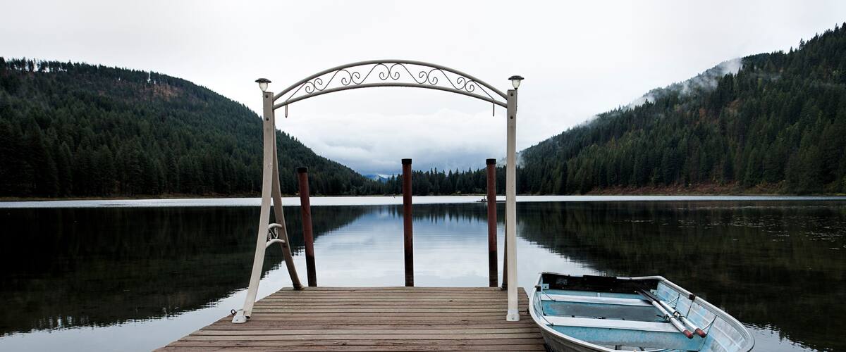 A wooden dock with a boat tied to it, surrounded by calm water and forested hills.