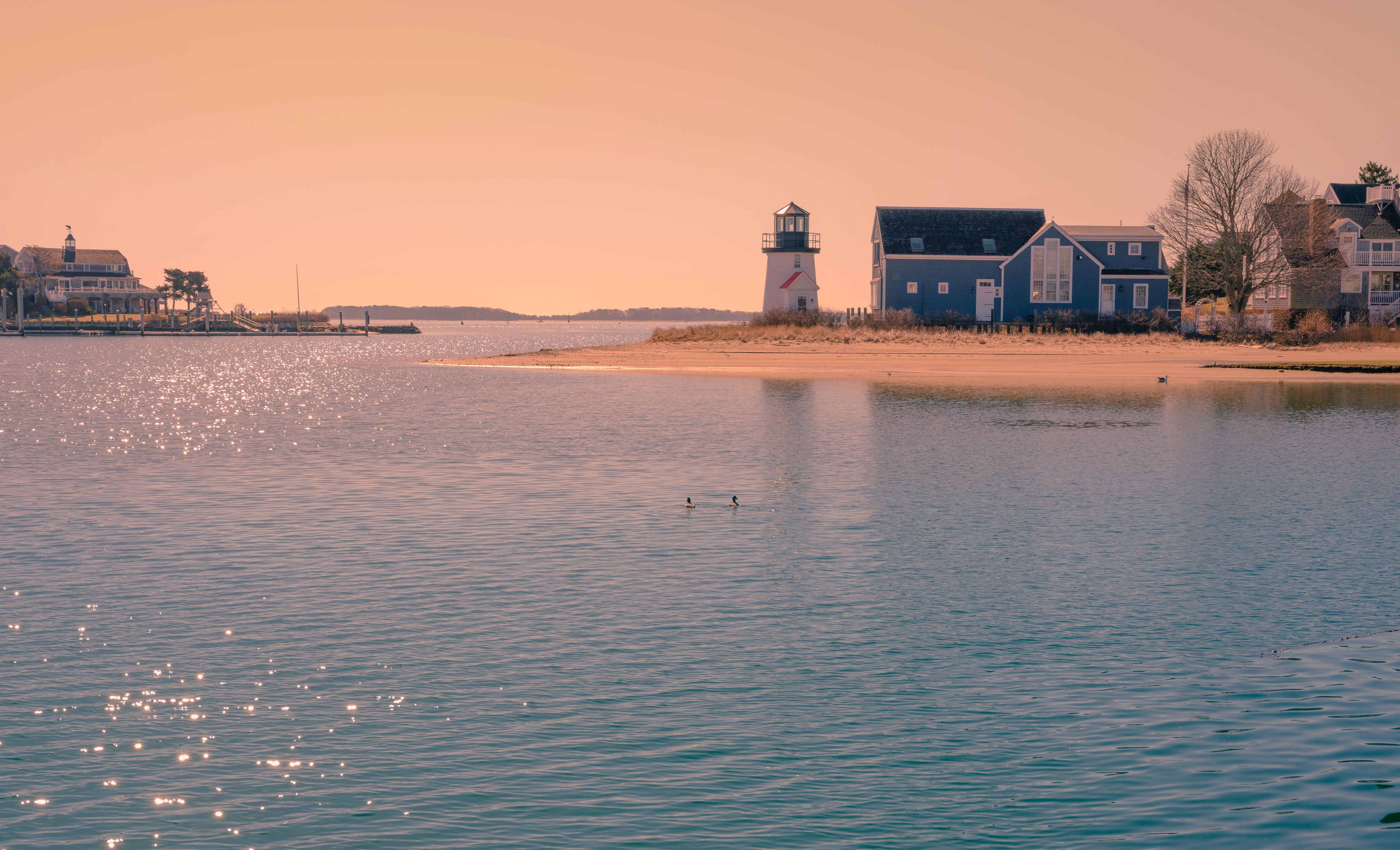 A calm waterfront with a lighthouse, a blue house, and a sandy beach.