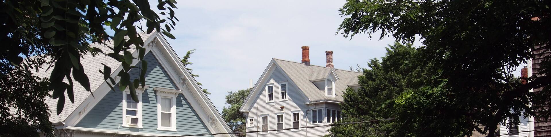 A row of historic houses with gabled roofs and white trim, surrounded by lush greenery and a well-maintained hedge.