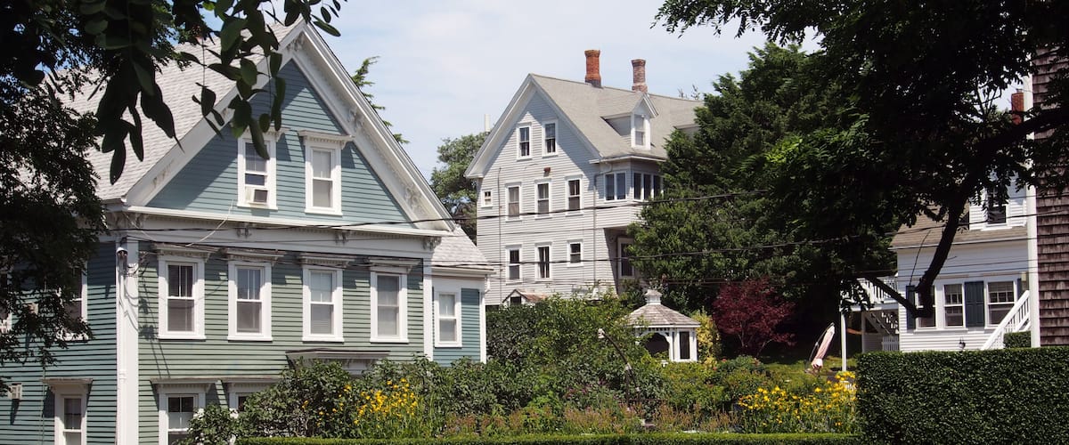 A row of historic houses with gabled roofs and white trim, surrounded by lush greenery and a well-maintained hedge.