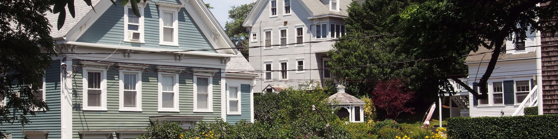 A row of historic houses with gabled roofs and white trim, surrounded by lush greenery and a well-maintained hedge.