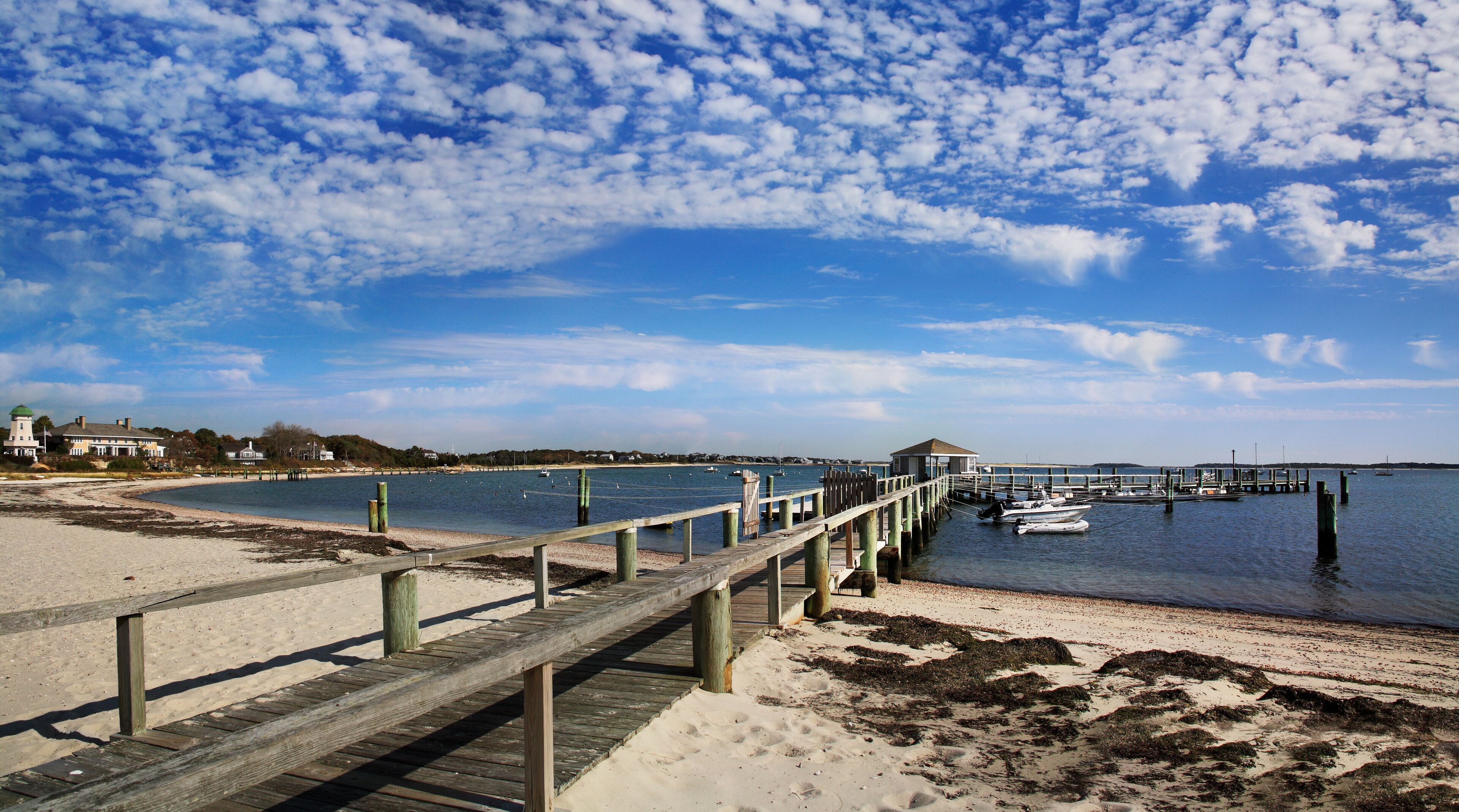A wooden pier extending into a calm body of water, with a sandy beach and a distant shoreline with buildings.