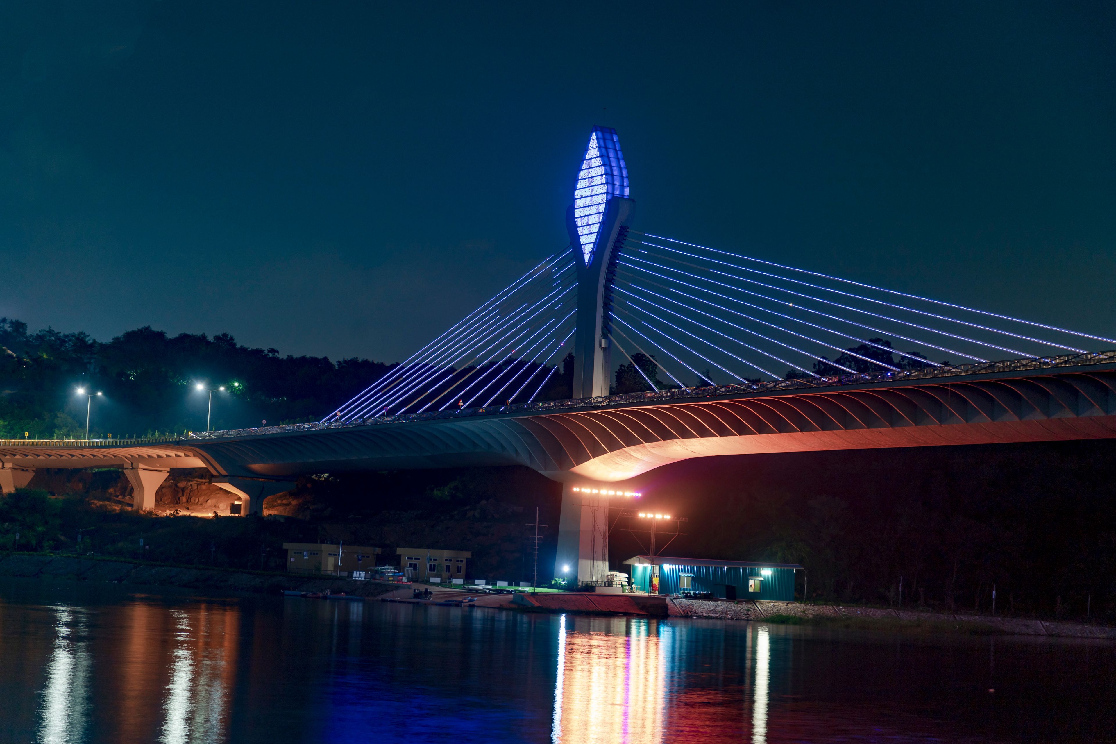 A modern bridge with a distinctive cable-stayed design, illuminated by blue lights against a night sky.