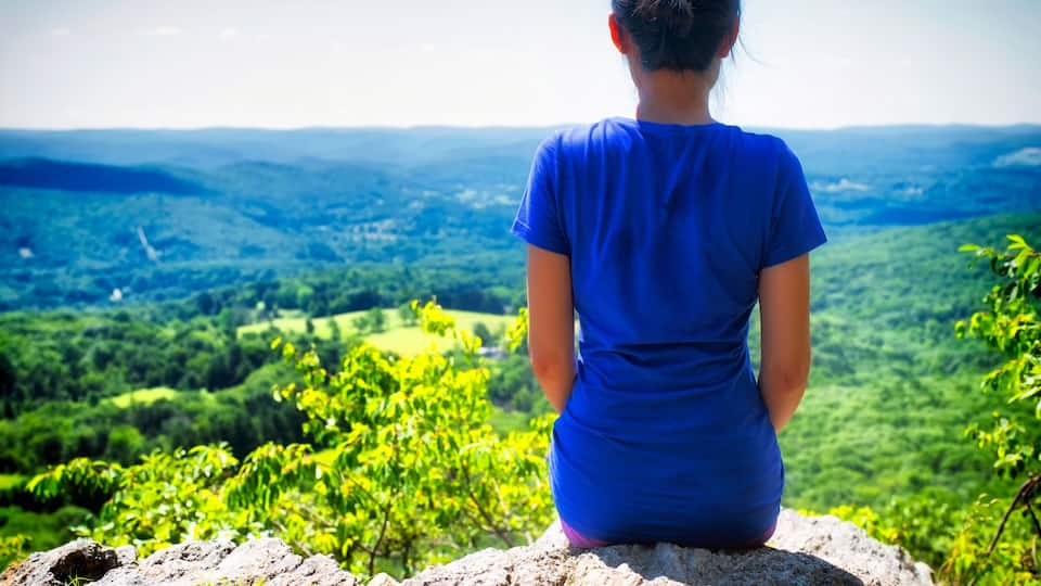 A person in a blue shirt sitting on a rock overlooking a green landscape.