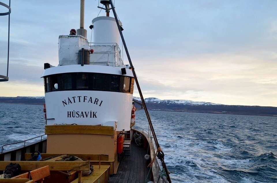 Un barco llamado Nattfari Husavik sobre una masa de agua, con montañas nevadas al fondo.