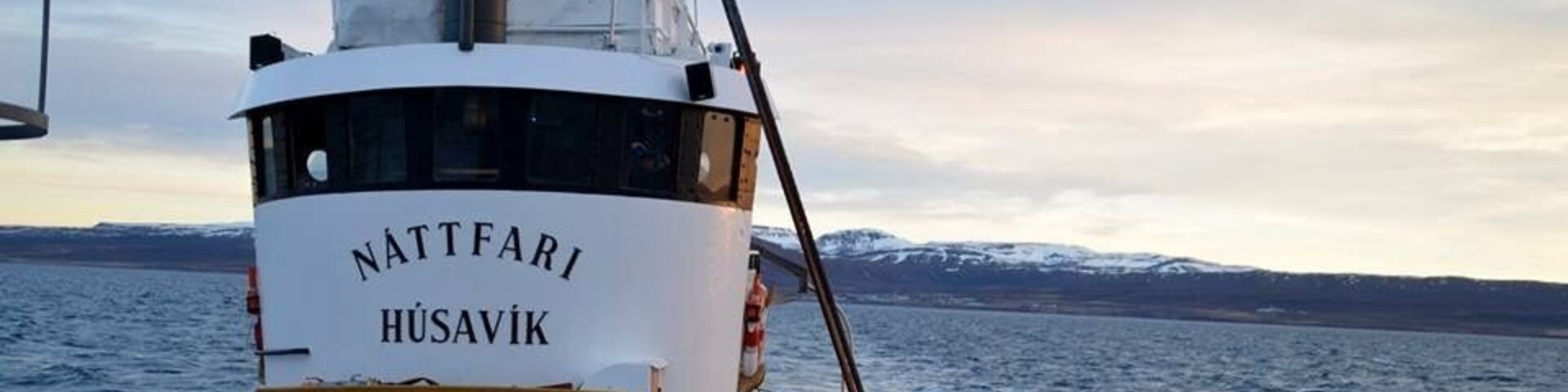 Un barco llamado Nattfari Husavik sobre una masa de agua, con montañas nevadas al fondo.