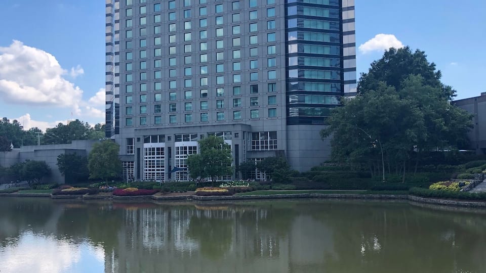 A modern high-rise building with a reflective glass facade, a calm water body in the foreground, and well-maintained greenery.