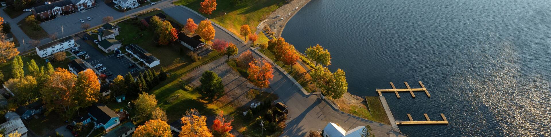 A stunning sunrise over Schroon Lake, NY, with warm light casting a soft glow on the calm water and surrounding autumn-colored forests and mountains.
