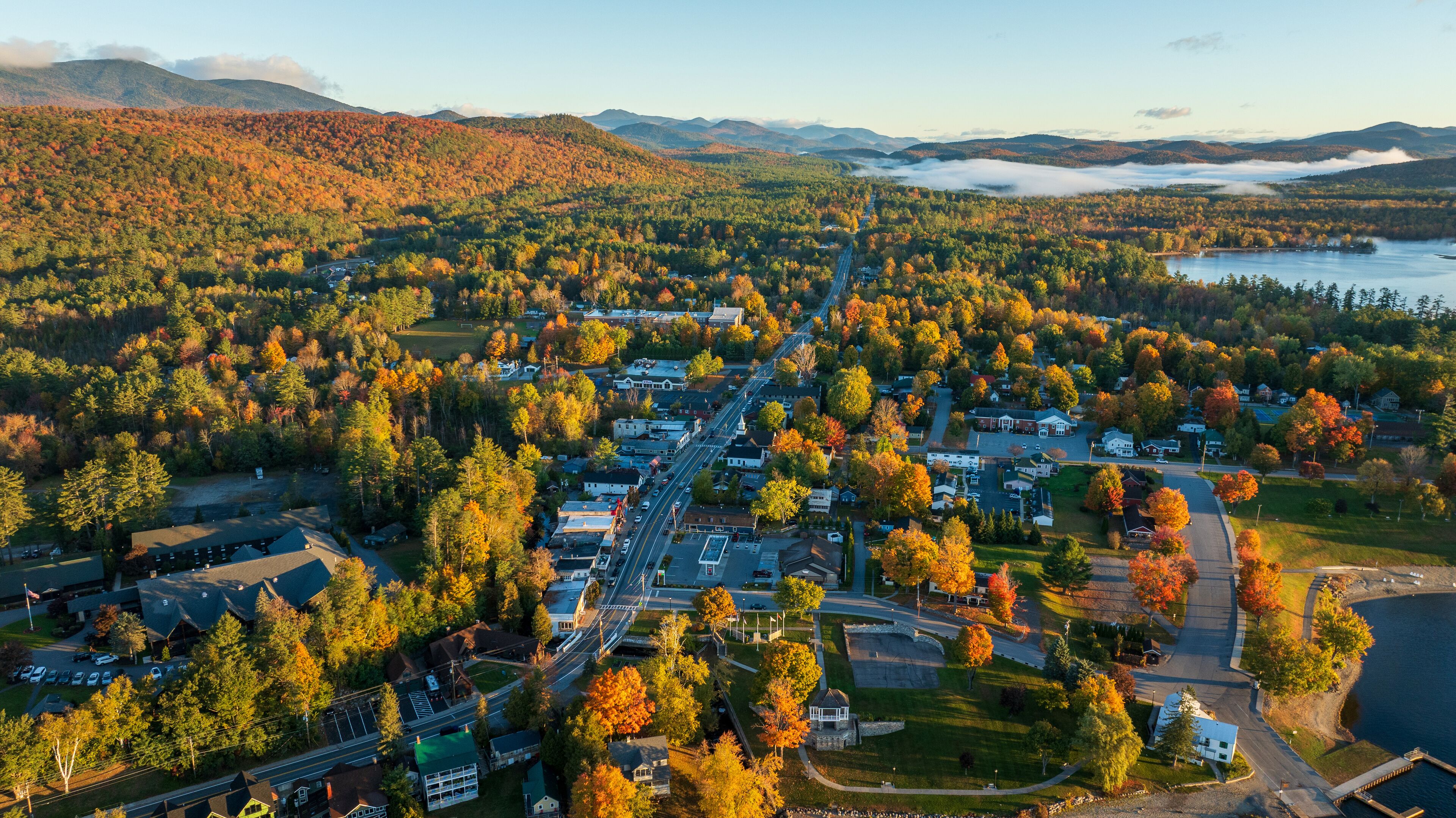 Aerial view of Schroon Lake, NY, with vibrant autumn colors, tree-lined streets, and surrounding Adirondack Mountains, showcasing the small town's scenic charm and peaceful lakeside setting.