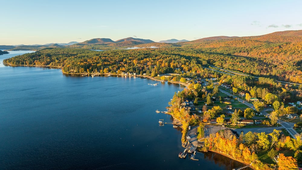 A stunning sunrise over Schroon Lake, NY, with warm light casting a soft glow on the calm water and surrounding autumn-colored forests and mountains.