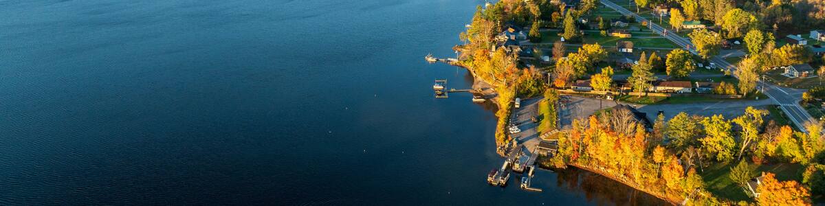 A stunning sunrise over Schroon Lake, NY, with warm light casting a soft glow on the calm water and surrounding autumn-colored forests and mountains.
